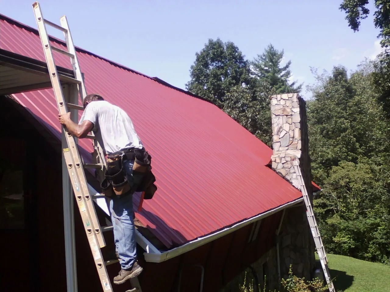 Roofer on a ladder beside a red metal roof and stone chimney — Soffit & Fascia Repair by Roofing Brothers in Horse Shoe